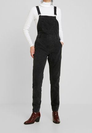 Person iført sort denim overalls, hvid rullekrave og brune hælede ankelstøvler står mod en ensfarvet lys grå baggrund.