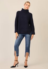 Maglione a collo alto a coste in navy con maniche lunghe, abbinato a jeans blu strappati con orlo decorato, e scarpe nere con tacco alto.