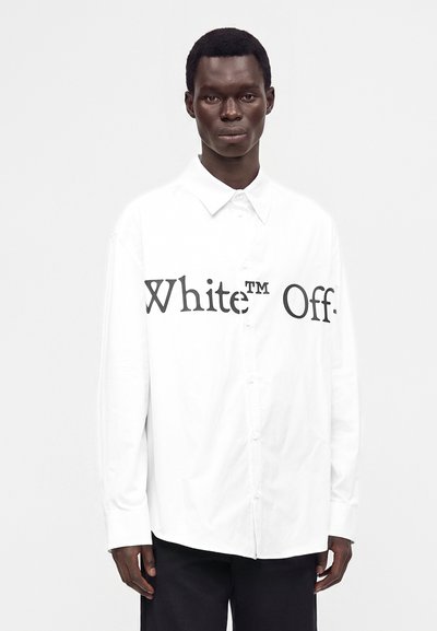 Млад мъж, носещ бяла риза с копчета с отпечатан текст "White™ Off." на гърдите, комбинирана с черен панталон.