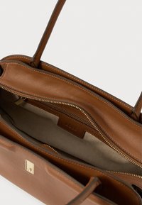 Bolsa de couro marrom com duas alças, topo aberto mostrando o forro interior bege e um bolso com fecho zip na parte interna.