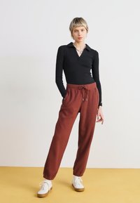 Schwarzes langärmliges Top mit kragenförmigem Ausschnitt, kombiniert mit rostfarbenen, gemusterten Hosen mit elastischem Bund und Kordelzug. Weiße Sneaker.