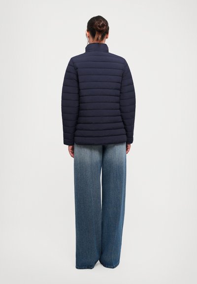 Woman with hair in bun wearing a navy blue quilted jacket and wide-leg faded blue jeans, standing facing away on a plain background.