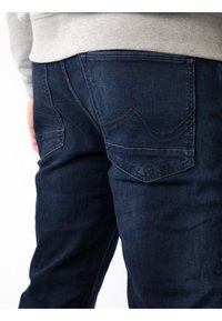 Jeans de denim azul oscuro con una textura suave, que presentan una costura curva distintiva en los bolsillos traseros y un diseño ajustado.