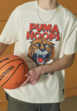 Person iført en "Puma Hoops" t-shirt med en brølende tiger grafik, der holder en orange basketball mærket "Training."
