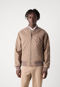 Tan bomberjack met een gladde, glanzende afwerking; ribgebreide manchetten en boord; vooraan een ritssluiting; gedragen over een witte bloes met kraag en beige broek.