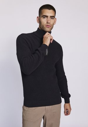 Maglione a collo alto a coste nere con maniche lunghe, caratterizzato da un zip decorativa sul colletto. Indossato sopra pantaloni beige, su uno sfondo chiaro.