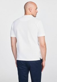 Biała koszulka polo z krótkim rękawem, klasycznym kołnierzykiem, o prostym kroju, z bocznymi rozcięciami oraz gładką bawełnianą fakturą. Noszona z ciemnymi spodniami.