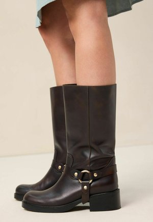 Braune Leder-Stiefel bis zum Knie mit runder Spitze, ausgestattet mit einem Riemen und dekorativen Beschlägen, auf einem Blockabsatz. Glatte Textur, minimalistisches Design.