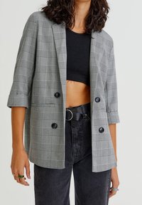 Blazer a quadri grigio con vestibilità rilassata, due bottoni neri e maniche accorciate. Indossato sopra un crop top nero e jeans neri a vita alta.