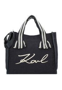Svart canvas-tygväska med en framträdande vit "Karl"-logotyp, svartvita randiga handtag och en avtagbar axelrem.