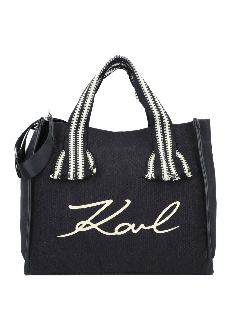 Svart canvas-tygväska med en framträdande vit "Karl"-logotyp, svartvita randiga handtag och en avtagbar axelrem.