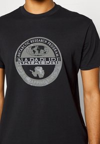 Camiseta negra de algodón con un gráfico circular gris que incluye los textos «Programa de Investigación Antártica» y «NAPAPIJR», además de un globo terráqueo y el contorno de la Antártida.