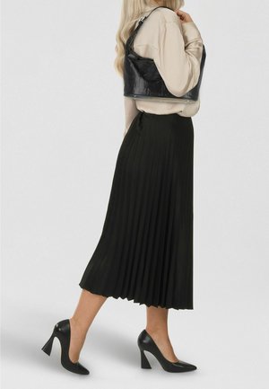 Zwarte geplooide midi-rok met een vloeiende textuur, gecombineerd met een lichtbeige blouse en zwarte pumps met hoge hakken, voorzien van een glanzende zwarte tas.