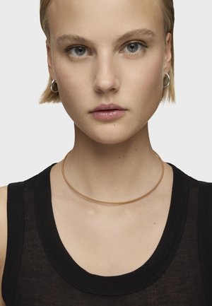 Metalowa choker na szyję o gładkim, smukłym wzornictwie w kolorze różowego złota, noszona z czarną, bezrękawnikową, prążkowaną bluzką. Styl minimalistyczny.