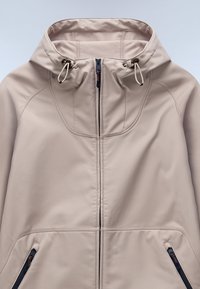 Beige, lätt jacka med hög krage, justerbar dragsko och två framfickor med dragkedja, som har slät tyg och minimala sömmar.