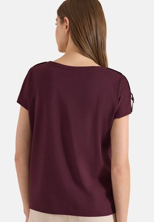 Femme aux longs cheveux bruns portant un haut violet foncé à manches courtes avec détail de bretelle sur l'épaule, tournée dos à la caméra.