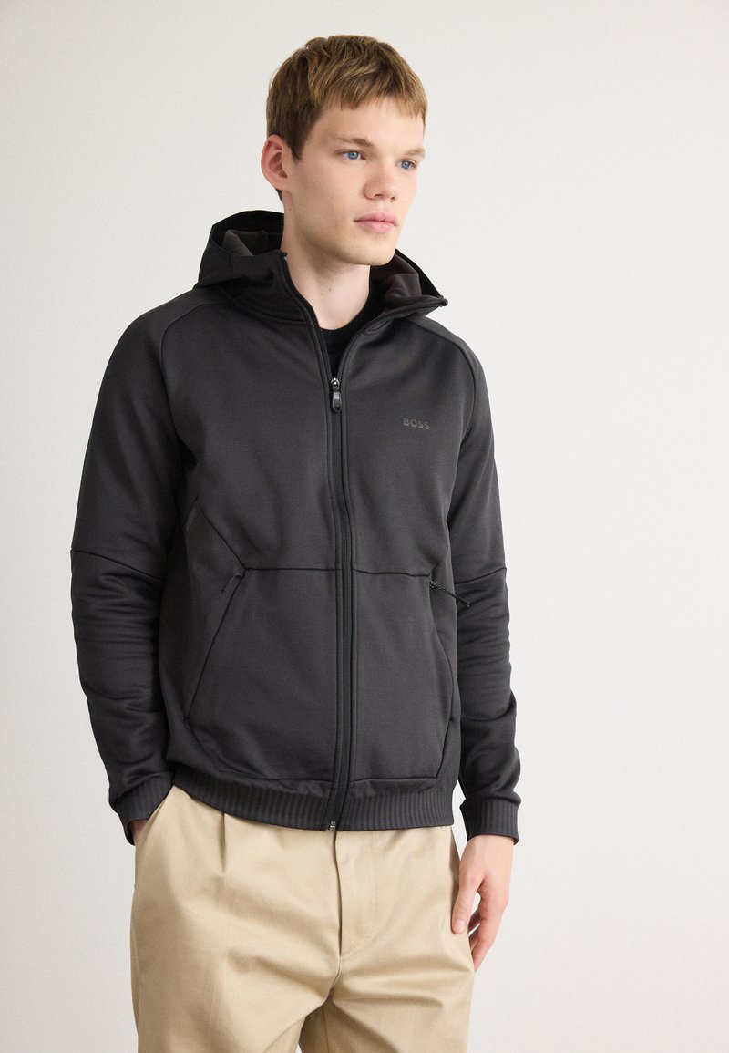 Siva zip-up kapuca z teksturirano tkanino, z visokim ovratnikom, dvema sprednjima žepoma in črnimi detajli. Nošena čez temno majico.