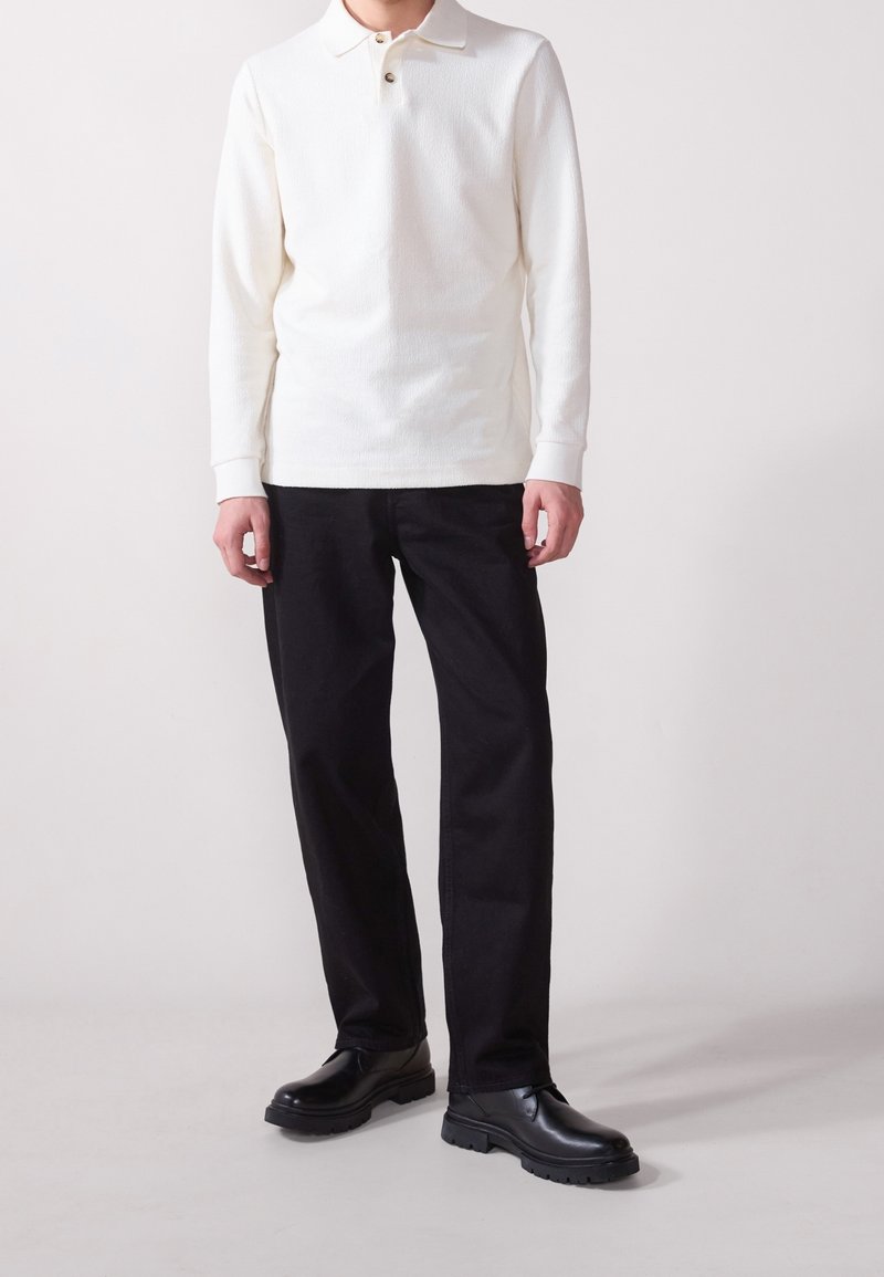 Homme portant un polo blanc à manches longues, un pantalon noir et des chaussures en cuir noires, debout devant un fond clair uni.