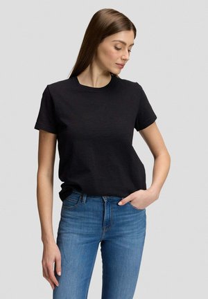 Junge Frau mit langen braunen Haaren, die ein schwarzes T-Shirt mit kurzen Ärmeln und blaue Jeans trägt, eine Hand in der Tasche, schaut nach unten und zur Seite.