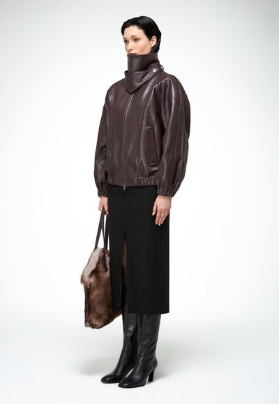Žena oblečená v tmavohnedej koženej bunde, čiernej midi sukni s predným rozparkom, čiernych čižmách na opätku a drží hnedú chlpatú kabelku.