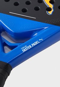 Blaues Padel-Racket mit strukturiertem Griff, mehreren Perforationen auf einer schwarzen Oberfläche und Markendetails in weißen und orangefarbenen Akzenten.