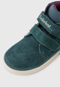 Grønn suedestøvel med borrelåsrem. Har et teksturert stofffôr i mørkeblått og en kremfarget gummisåle. Timberland-merke på remmen.