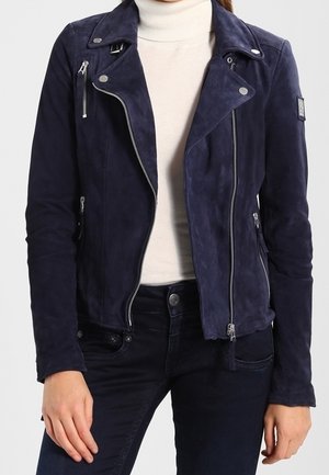 Persona che indossa una giacca da motociclista in camoscio blu navy con cerniere argentate sopra un dolcevita bianco e jeans scuri.