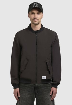 Chaqueta bomber negra con cremallera frontal, puños y dobladillo de canalé, dos bolsillos laterales y un parche de logotipo en la parte inferior. Llevada con una gorra negra.