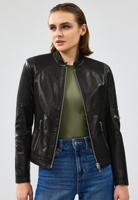 Chaqueta de cuero negra con acentos acanalados en los hombros, cierre frontal con cremallera y bolsillos laterales. Lleva puesta una blusa verde y unos vaqueros de tiro alto azules.