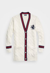 Stickad kavaj i krämfärg med kabelmönster, ribbade manschetter och marinblå-röd kant. Har två framfickor och en broderad kunglig krans.