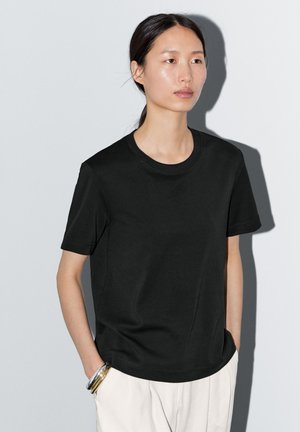 T-shirt nera in cotone con scollatura rotonda e maniche corte, caratterizzata da una vestibilità comoda e una superficie liscia. Abbinata a pantaloni di colore chiaro.