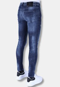 Strakke spijkerbroek in donkerblauw denim met vervaagde plekken, voorzien van vijf zakken en een zwart merklogo tailleband, gecombineerd met witte sneakers.