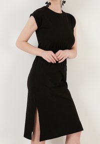 Kobieta w czarnej, midi sukience z krótkim rękawem oraz rozcięciem z boku, w czarnych kwiatowych kolczykach, stojąca przy białej ścianie.