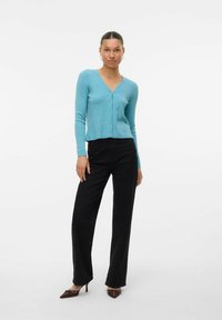 Cardigan côtelé bleu clair avec un décolleté en V profond et une fermeture à boutons, associé à un pantalon large noir et des mules à talons marron.
