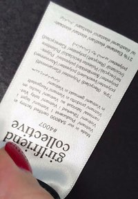 Main tenant une étiquette de vêtement blanche avec la composition détaillée du tissu et le texte de la marque « giftfriend collective » sur un fond sombre.