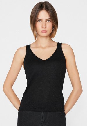 Giovane donna con capelli castani corti che indossa un top nero senza maniche con scollo a V e pantaloni scuri, in piedi davanti a uno sfondo bianco uniforme.
