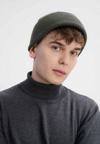 Donkergrijze coltrui met een ribbelstructuur, gecombineerd met een groene gebreide muts. Het model heeft kort, tousled haar en een neutrale uitdrukking.