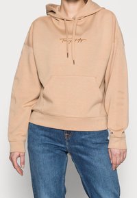 Beige hoodie tillverkad av mjukt material, med en framficka, justerbar dragsko i huvan och broderad logotyp i kontrasterande färg.