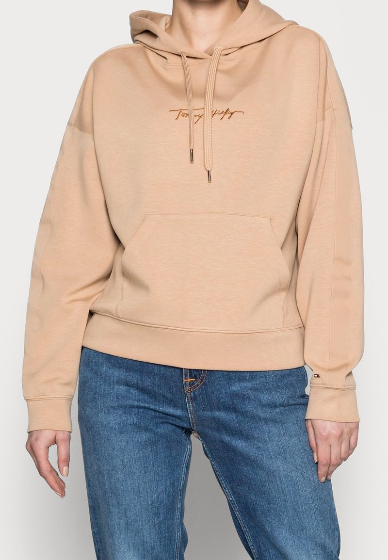 Beige hoodie tillverkad av mjukt material, med en framficka, justerbar dragsko i huvan och broderad logotyp i kontrasterande färg.