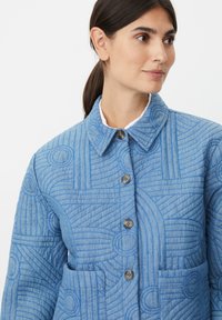 Blå quiltet jakke med krave, der har cirkulære og kantede mønstre, knaplukning foran og en lomme med lap på venstre side.