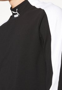 Czarna bluzka z długim rękawem z białymi akcentami na ramionach; ma wysoki kołnierz i logo Puma na szyi. Wykonana z miękkiego materiału.