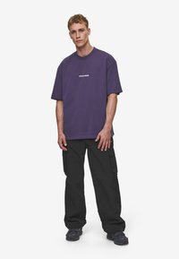 Violeta oversize t-kreklis ar baltu logo, savienots ar melniem cargo biksēm ar sānu kabatām. Modelis valkā tumšus sporta apavus.