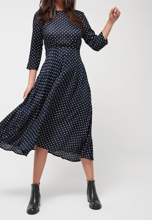 Navy Kleid mit weißen Punkten, Dreiviertelärmeln, eng am Oberkörper und mit weit ausgestellt Rock in knielanger Länge. Kombiniert mit schwarzen Knöchelstiefeln.