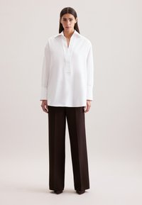 Camicia bianca a maniche lunghe con colletto e patta parziale, abbinata a pantaloni larghi a vita alta in tessuto morbido di colore marrone.