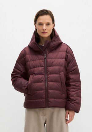 Vrouw met kort bruin haar die een donkerrode doorgestikte pufferjas met capuchon en beige broek draagt tegen een effen lichte achtergrond.