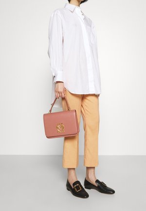 Persoon gekleed in een witte oversized blouse, beige cropped broek, zwarte loafers met gouden gesp, en een roze handtas met gouden logo, tegen een eenvoudige achtergrond.