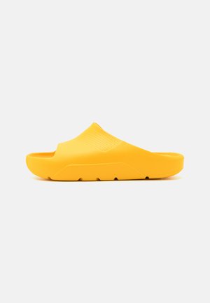 Zueco amarillo hecho de goma suave, con parte superior perforada, plantilla contorneada y suela texturizada para mayor agarre.