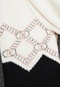 Sciarpa lavorata a maglia con una parte superiore bianca decorata da un motivo a diamante accentuato da borchie argentate e rosa oro, che si trasforma in una parte inferiore nera.