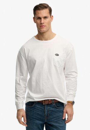 Man wearing white long-sleeve shirt with small logo on chest pocket, blue jeans, brown leather belt, and wristwatch on left wrist.