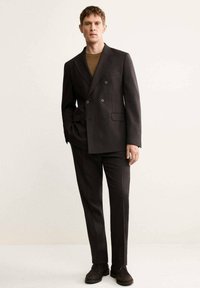 Blazer doppiopetto marrone scuro abbinato a pantaloni coordinati. Tessuto testurizzato, vestibilità slim e dettagli con bottoni neri. Scarpe in suede marroni.